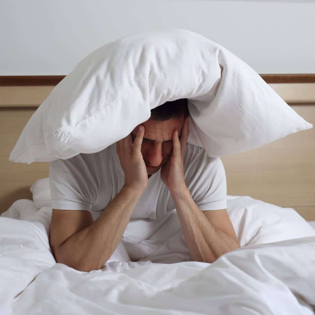 Man met slaapproblemen zit rechtop in bed met kussen op hoofd