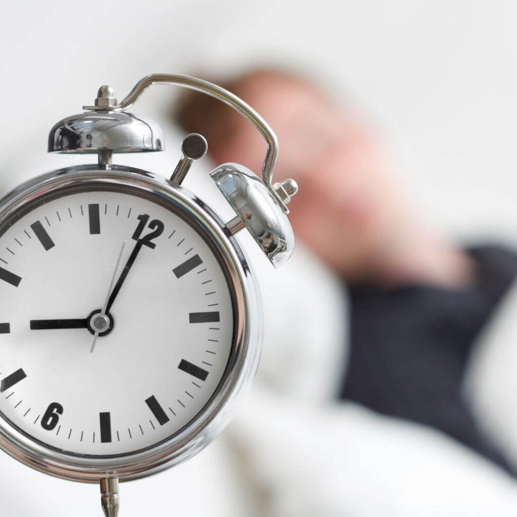 Man in bed met klok op voorgrond: hoeveel slaap nodig?