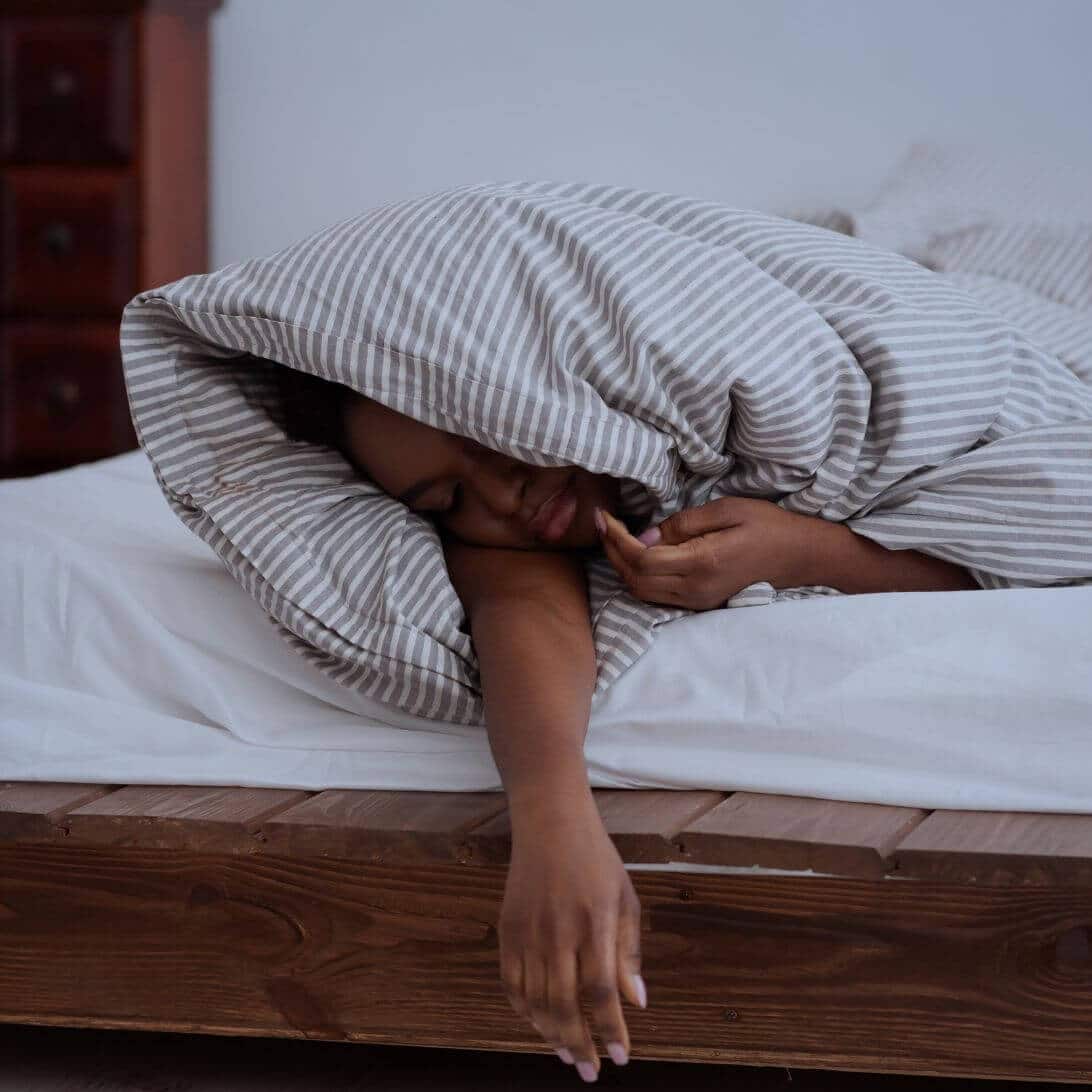 Vrouw ligt met haar hoofd aan de voetenkant in bed, onder de dekens en met een arm buiten het bed