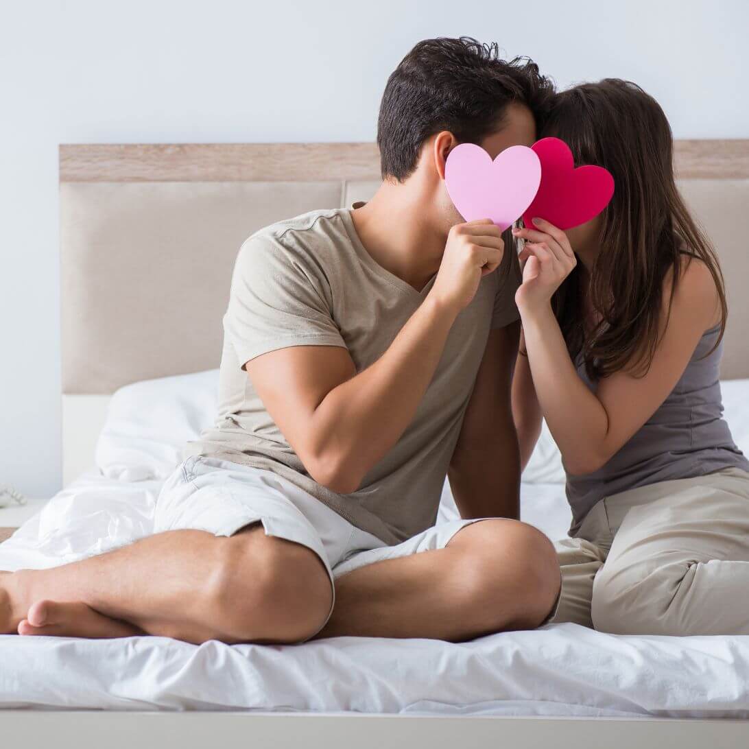Man en vrouw zitten op bed en hebben een roze en rode hartje voor hun gezicht